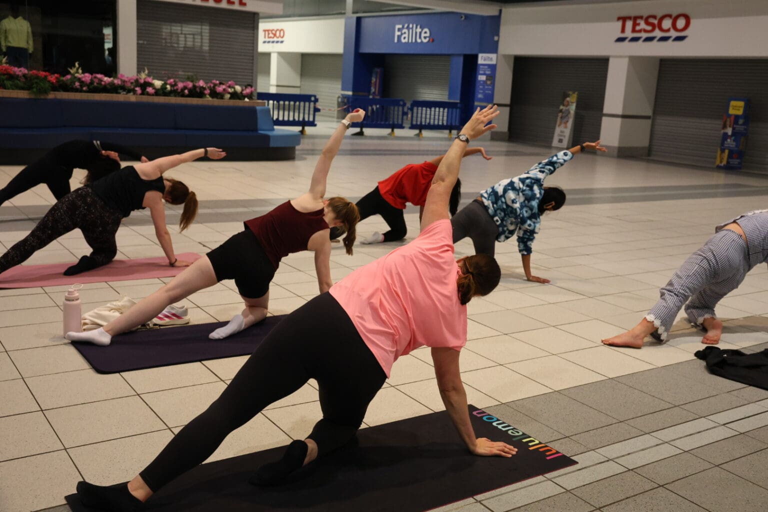 A yoga class in side angle pose on mats in the Crescent Shopping Centre concourse, with Tesco visible in the background.