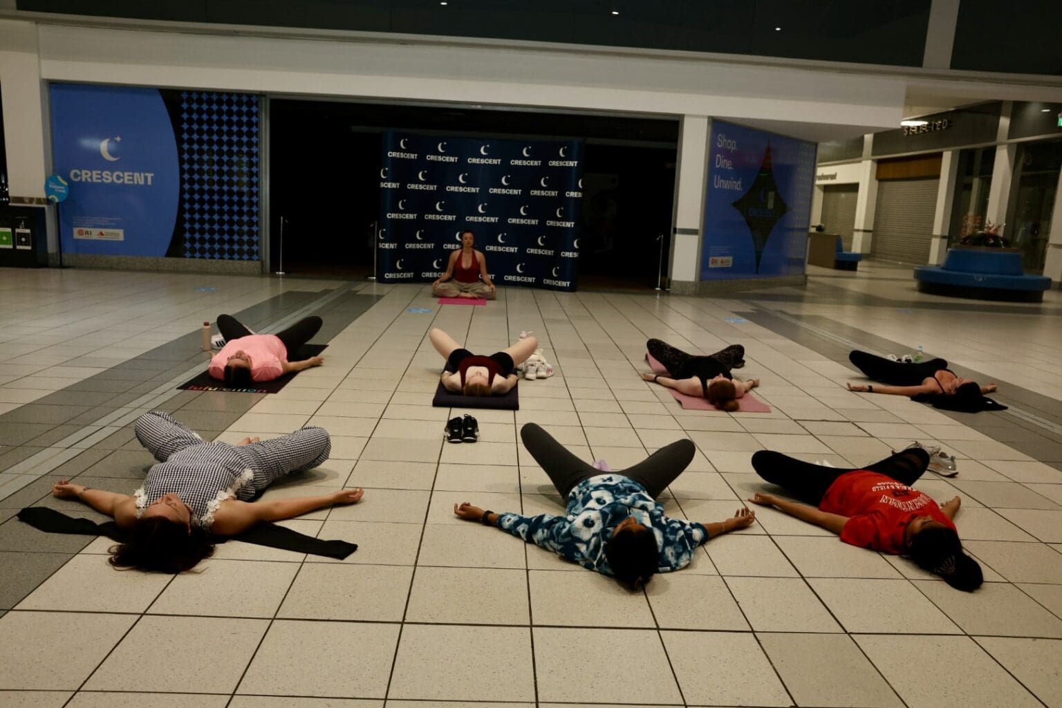 A yoga class lying in savasana on mats in the Crescent Shopping Centre concourse, with the instructor seated at the front.