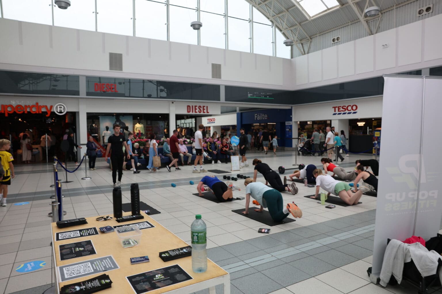 A fitness class on mats in the main atrium at Crescent Shopping Centre, with shoppers walking past and the glass roof above.